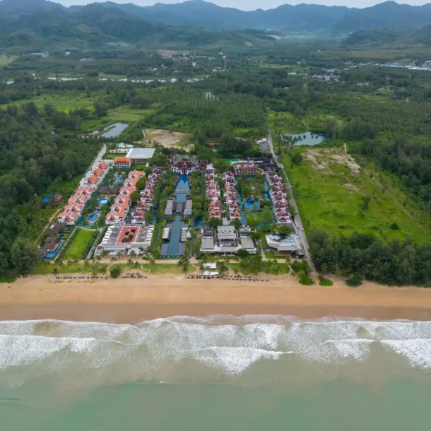 An aerial view of a large beach resort complex with a swimming pool and buildings, juxtaposed with a mountain range in the background, perfectly illustrating the choice between Beach Resorts vs Mountain Resorts.